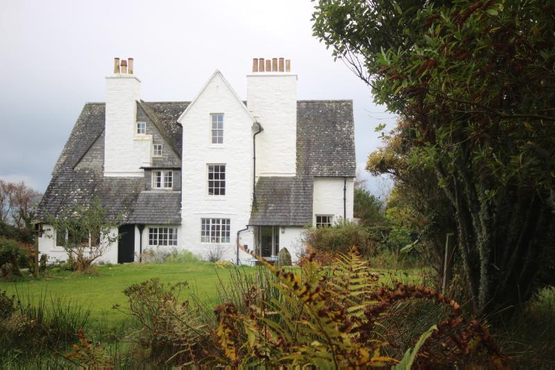 Dun A Bhuilg House, rear elevation, looking east during daytime with white sky and trees in front of the house. 