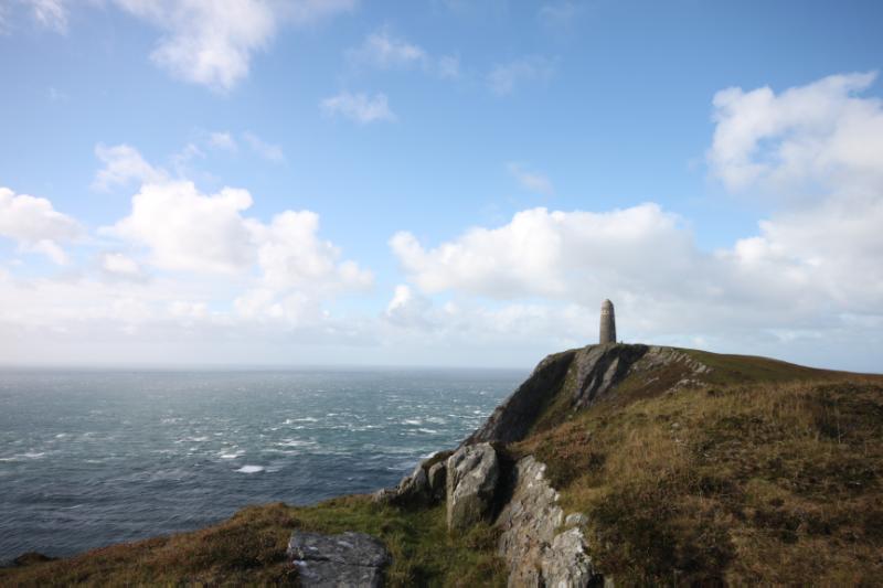 The American Monument, located on the cliff top at the southern end of Mull of Oa. Overlooking looking the site of the wreck of the SS Tuscania. Looking west, during daytime, on a clear day with patchy blue sky and stormy sea.