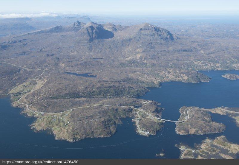 Oblique aerial view of Drochaid a’ Chaolais Chumhaing / Kylesku Bridge, with Quinag beyond, looking soutwest, during daytime with blue sky.