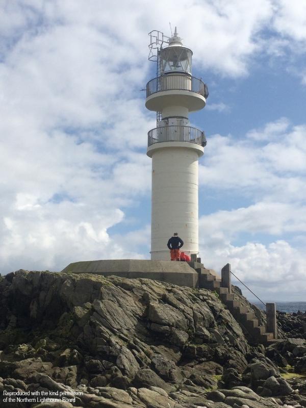 Ve Skerries Lighthouse, principal elevation, during daytime, with blue sky and clouds. Rocks in the foreground and the sea in the background. A person stands at the base of the lighthouse.  