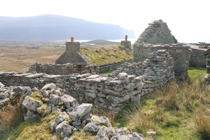 Rear of cottage with remains of farm buildings in foreground, looking southeast. Rackwick bay and seacliffs in the distance, on a dry day with white sky
