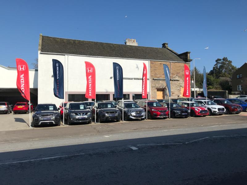 South elevation of former volunteer drill hall with cars for sale and banners in foreground, looking north, during daytime on clear day with blue sky.