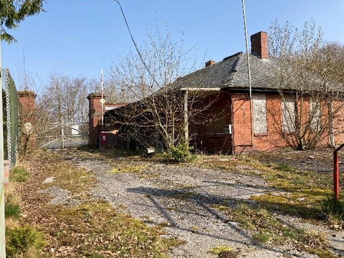 East guardhouse and pair of gatepiers at the former entrance to Eastriggs factory, rear elevation, looking northeast, with trees and vegetation growth in foreground, during daytime, on clear day with blue sky.