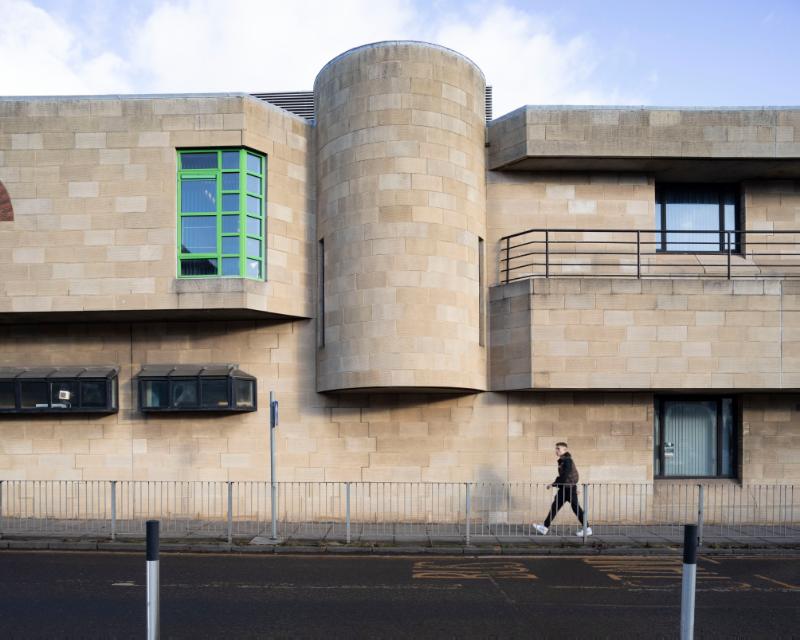 Tollcross Fire Station, west elevation detail, looking east, on a clear day with cloudy blue sky and a person walking past. 