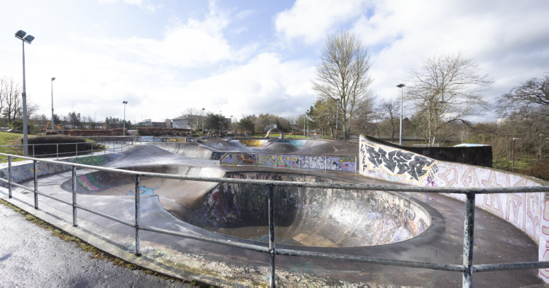 Livingston skatepark, general view from north, railings in foreground, trees and streetlight in background, blue-white sky. 